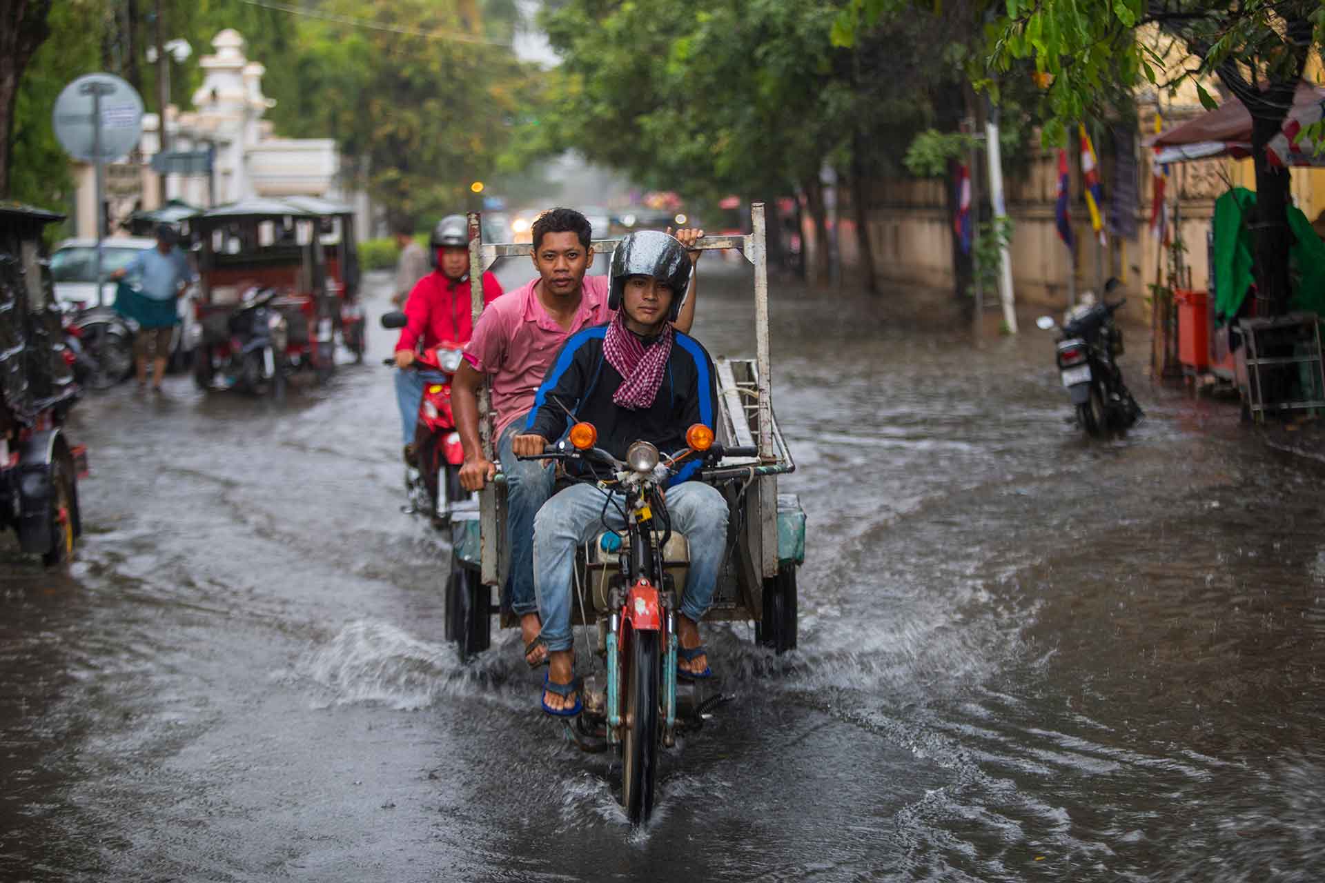 Two motorcyclists riding through a flooded urban street in Southeast Asia during heavy rainfall